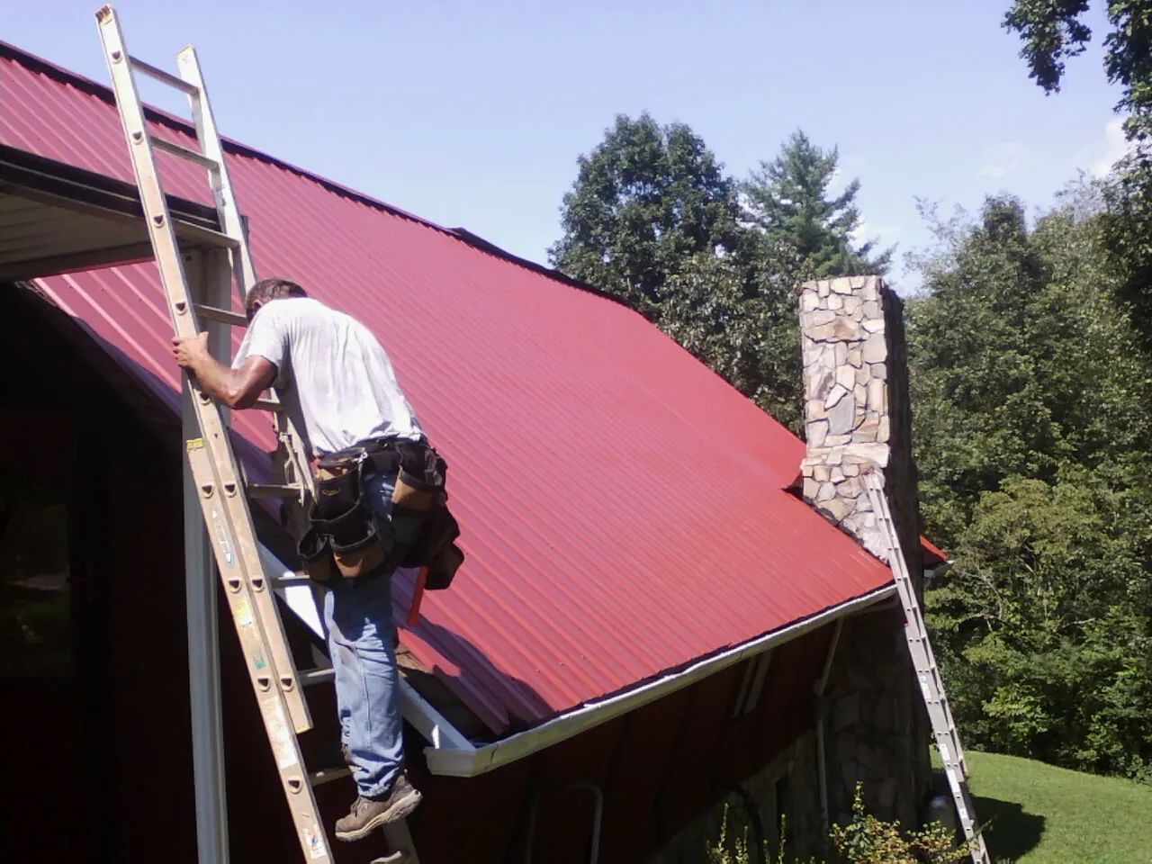 Roofer on a ladder beside a red metal roof and stone chimney — Soffit & Fascia Repair by Roofing Brothers in Hamburg